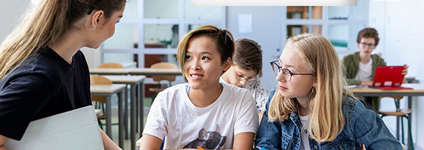 Två ungdomar som sitter i bänkarna bredvid varandra i klassrummet och som pratar med läraren