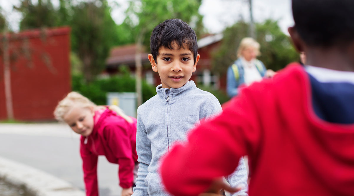 Pojke på en skolgård, tittar in i kameran, två skolkamrater vid sidan om honom