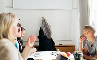 En person skriver på en whiteboard-tavla medan kollegor sitter runt ett bord