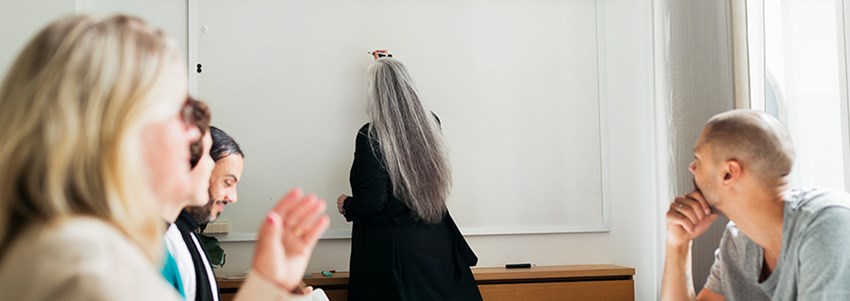 En person skriver på en whiteboard-tavla medan kollegor sitter runt ett bord